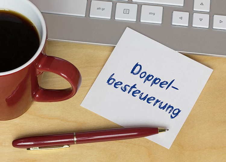 Zettel mit der Aufschrift „Doppelbesteuerung“. Daneben eine Tastatur, ein Kugelschreiber und eine Tasse Kaffee.