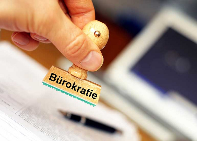 Hand hält Stempel mit Aufschrift "Bürokratie". Im Hintergrund ist ein Schreibtisch/Arbeitsplatz zu sehen.