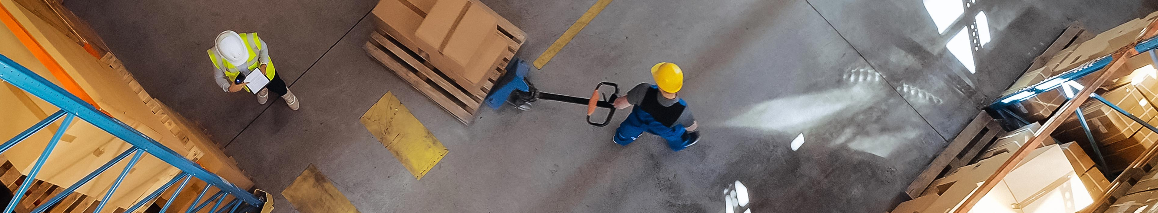 A warehouse worker moves boxes on a Euro pallet with a forklift truck. Another person checks data on a clipboard next to a shelf.