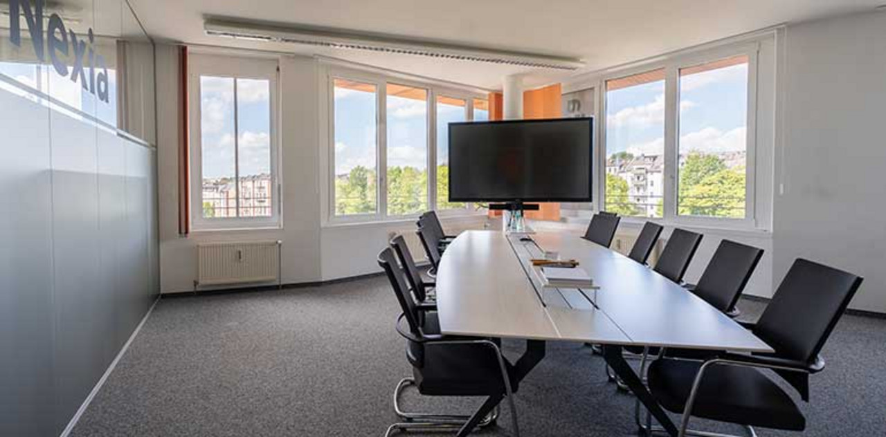 Nexia GmbH | Chemnitz Der Blick fällt in einen leeren Meetingraum mit einem Tisch und zehn Stühlen. Zudem ist ein Fernseher zu sehen. Der Blick geht über eine Fensterfront nach draußen.
