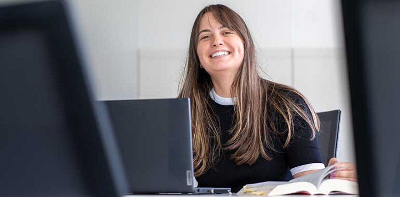 Weiterbildung Person sitzt mit einem Buch an ihrem Arbeitsplatz und lacht.
