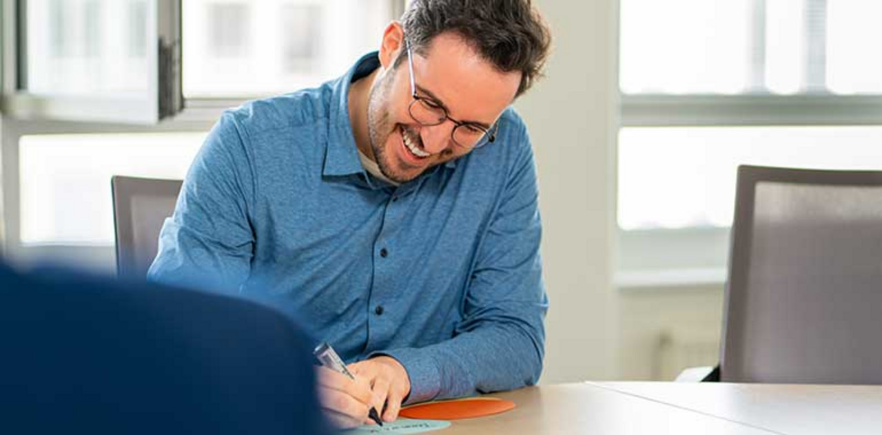 Eine Person sitzt lachend an einem großen Tisch und schreibt etwas auf einen Zettel. Learning on-the Job
