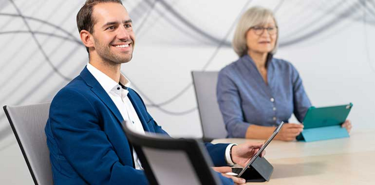 Nexia Hub An einem großen Tisch sitzen zwei Personen mit einem Tablet und schauen nach vorne.