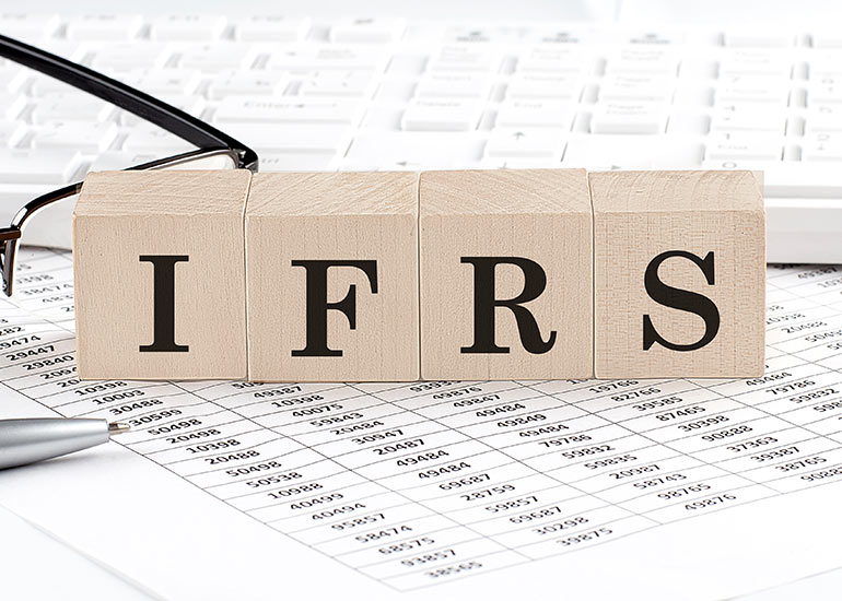 Bauklötze, die die Buchstaben 'IFRS' bilden. Diese sind auf Blättern mit Zahlentabellen und einer Lesebrille angeordnet. Jeder Buchstabe steht auf einem eigenen Klotz, die zusammen den Begriff IFRS (International Financial Reporting Standards) darstellen.