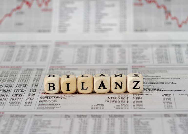 Auf Würfeln steht "Bilanz". Jeder Buchstabe steht auf einem eigenen Würfel. Im Hintergrund ist der Börsenteil einer Zeitung zu sehen.