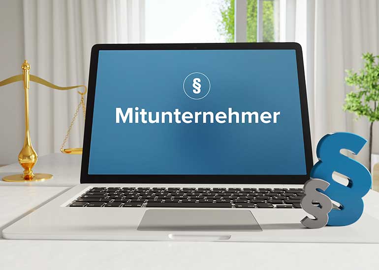 Auf einem Schreibtisch steht ein Laptop, auf dessen Bildschirm ein Paragraphenzeichen und das Wort "Mitunternehmer" zu sehen sind. Im Hintergrund befindet sich eine Richterwaage und daneben zwei weitere Paragraphenzeichen.