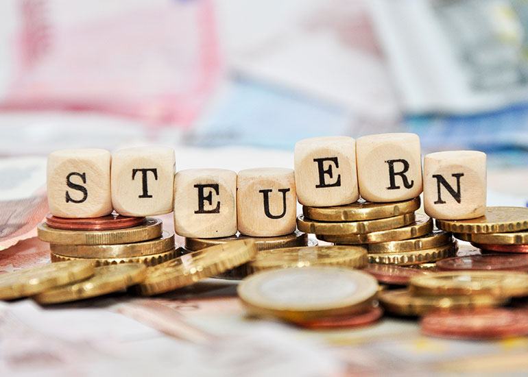 Der Begriff "Steuern" ist auf Würfeln geschrieben. Jeder Buchstabe ist auf einem einzelnen Würfel abgebildet. Alle Würfel stehen jeweils auf einem Turm aus Münzen.