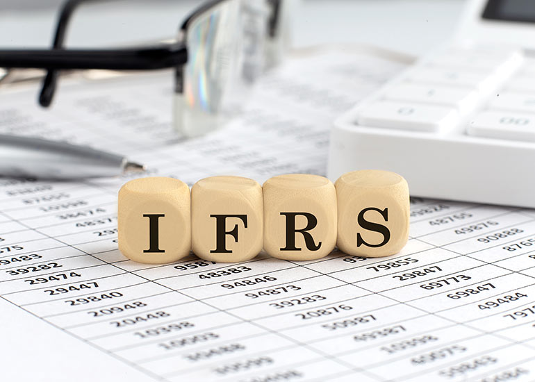 Auf einem Tisch liegen Würfel, auf denen „IFRS“ für „International Financial Reporting Standards“ steht. Jeder Buchstabe steht auf einem Würfel. Unter den Würfeln liegt ein Blatt mit Zahlen. Im Hintergrund liegen eine Brille und ein Taschenrechner.